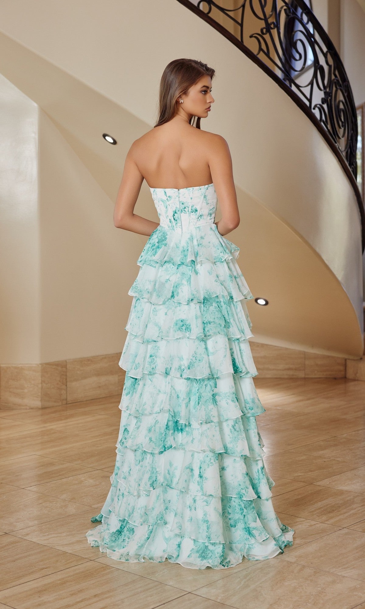 A woman stands indoors facing away, wearing the Nox Anabel T1865—a strapless floral chiffon long prom dress with green marble patterns and an A-line ruffle skirt.